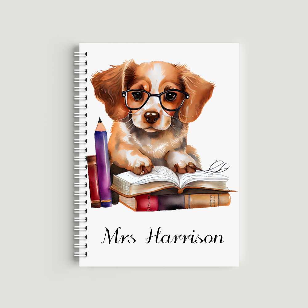 A spiral-bound A5 notebook with a watercolor illustration of a brown and white puppy wearing black glasses, sitting behind a stack of books with a pencil. The name "Mrs Harrison" is printed at the bottom in serif font.