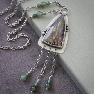 A sterling silver pendant shaped like an arrowhead, with a moss agate gemstone and round moss agate beads on a silver chain. Resting on a green box with a purple suede background.
