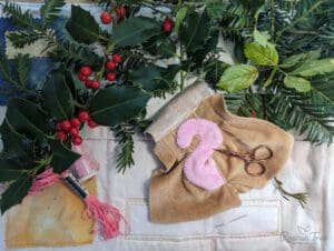 holly leaves and berries surrounding brown mouse doll tail appliqué work ready for sewing with skein of pink embroidery thread placed on left side next to spool of pink sewing thread