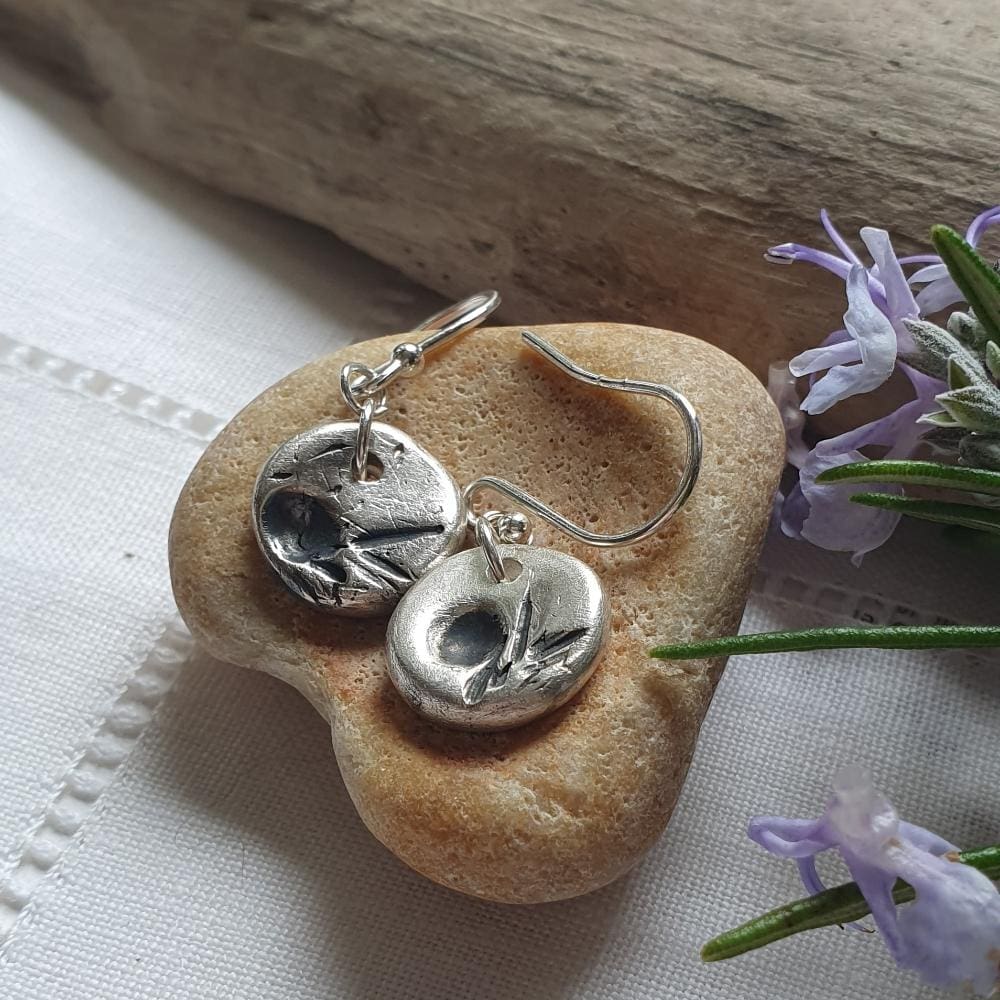 A pair of dainty silver disc earrings, each with a slightly different imprint of a juniper plant.