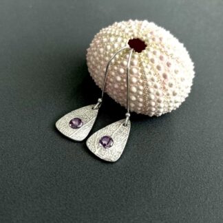 À pair of fine silver drop earrings with a amythst stone set in the centre of Rachel one. They have sterling silver ear wires and displayed on a dark grey background with a pink sea shell above them.