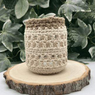 a little glass jar covered in a lacey crochet pattern with jute finish round the neck of the jar. It's standing on a log slice and there is greenery in the background