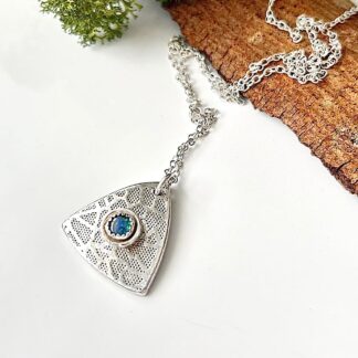 A fine silver triangular shaped pendant with a 5mm. opal stone in the centre. It has a textured pattern and a sterling silver chain, it is displayed on a white surface with the chain resting on some wood bark.