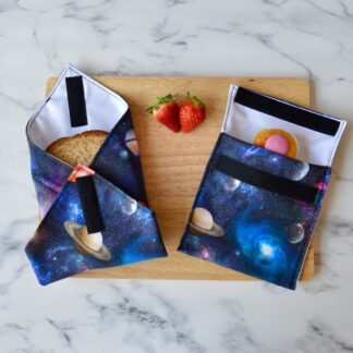 Planets space-themed sandwich wrap and snack bag on a wooden board. The fabric is navy with planets and star clusters. A sandwich is partially wrapped in the sandwich wrap, a cupcake is inside the snack bag, and fresh strawberries are placed on the board.