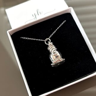 A fine silver poppy sede head with a sterling silver chain. The pendant is displayed on a brown glass background with a small white presentation box in the top left corner.