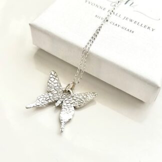 A fine silver pendant in the shape of a small butterfly, the pendant has a sterling silver chain. It is displayed on a black velvet pad inside a small white presentation box.