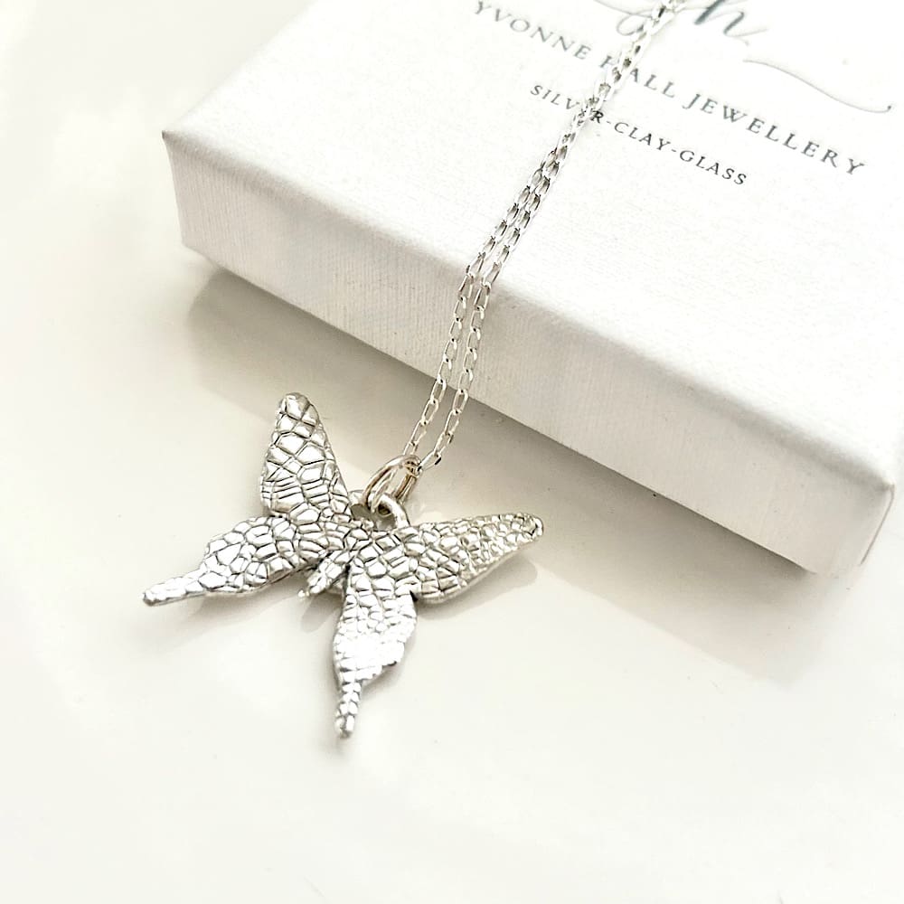 A fine silver pendant in the shape of a small butterfly, the pendant has a sterling silver chain. It is displayed on a black velvet pad inside a small white presentation box.