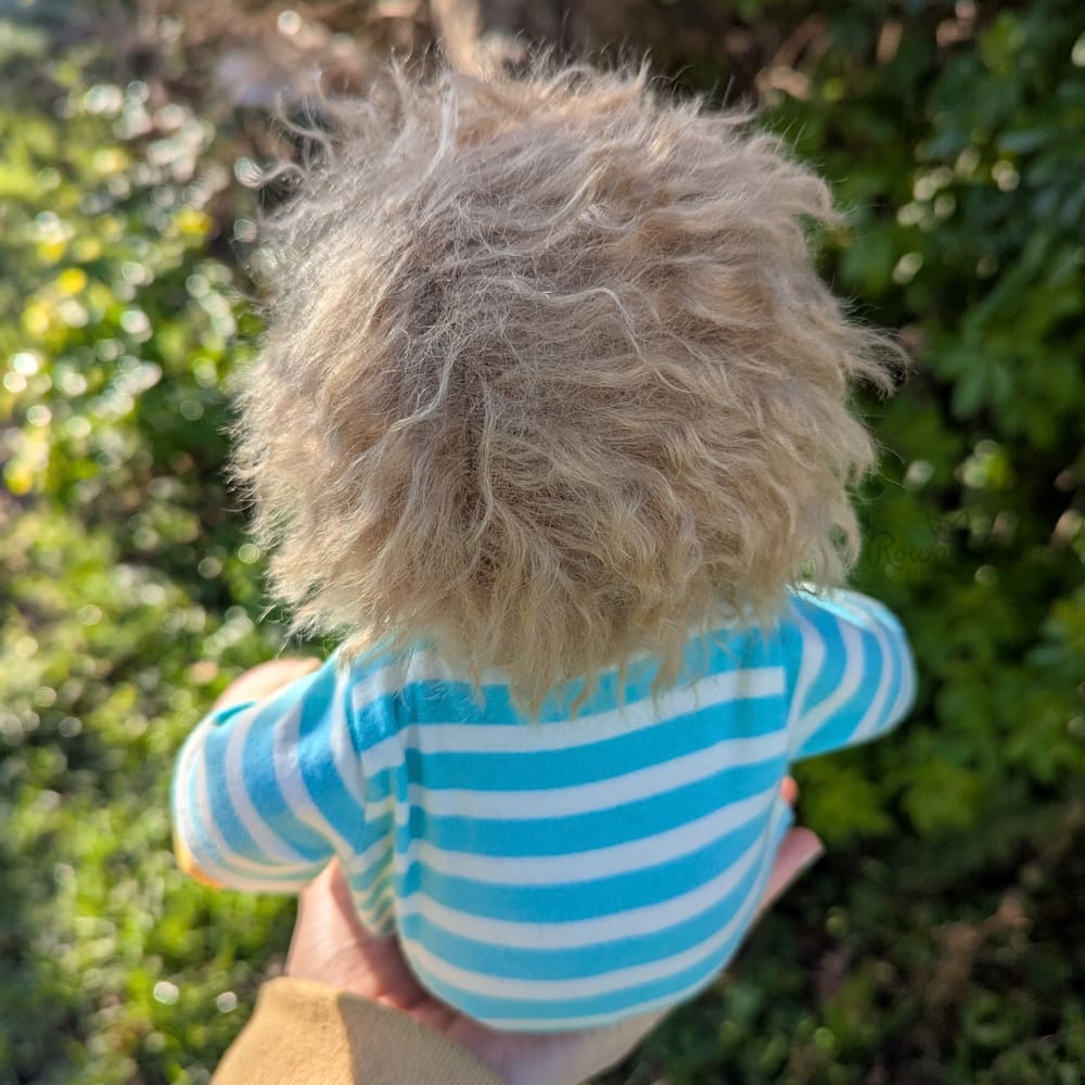 handmade fabric baby doll with sandy brown hair dressed in onesie in a garden background sat on a hand showing rear of body and head