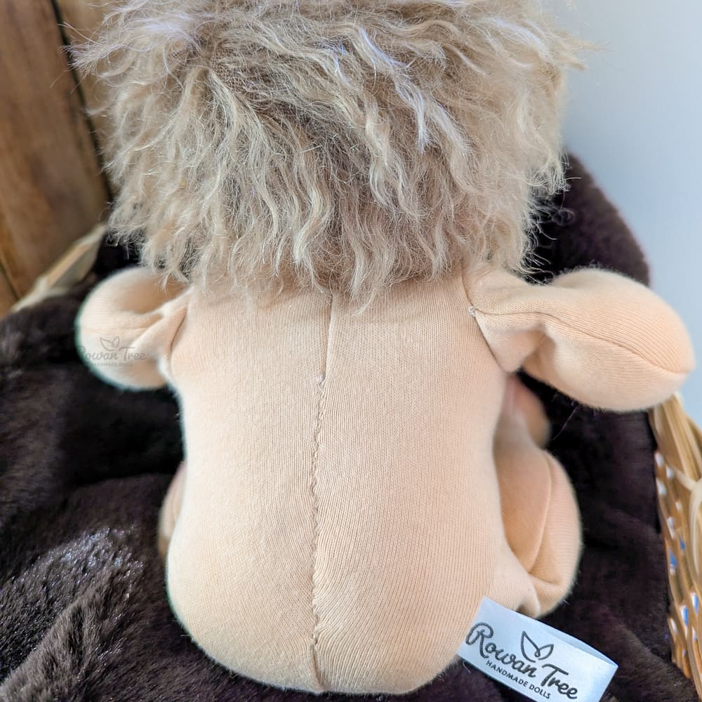 handmade fabric baby doll with brown curly hair seated in basket showing back view with Rowan Tree Doll label