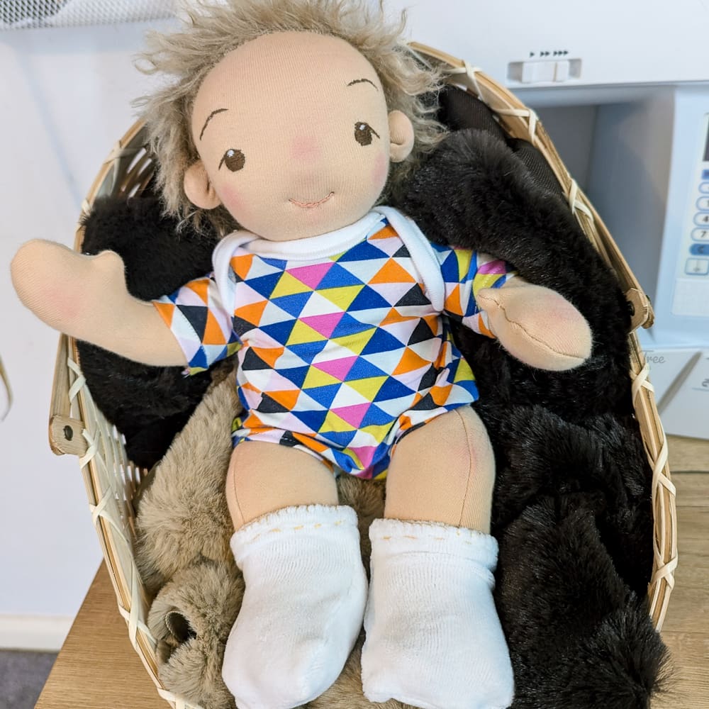 handmade fabric baby doll with sandy brown hair and brown eyes in a colourful bodysuit and white socks waving from a basket