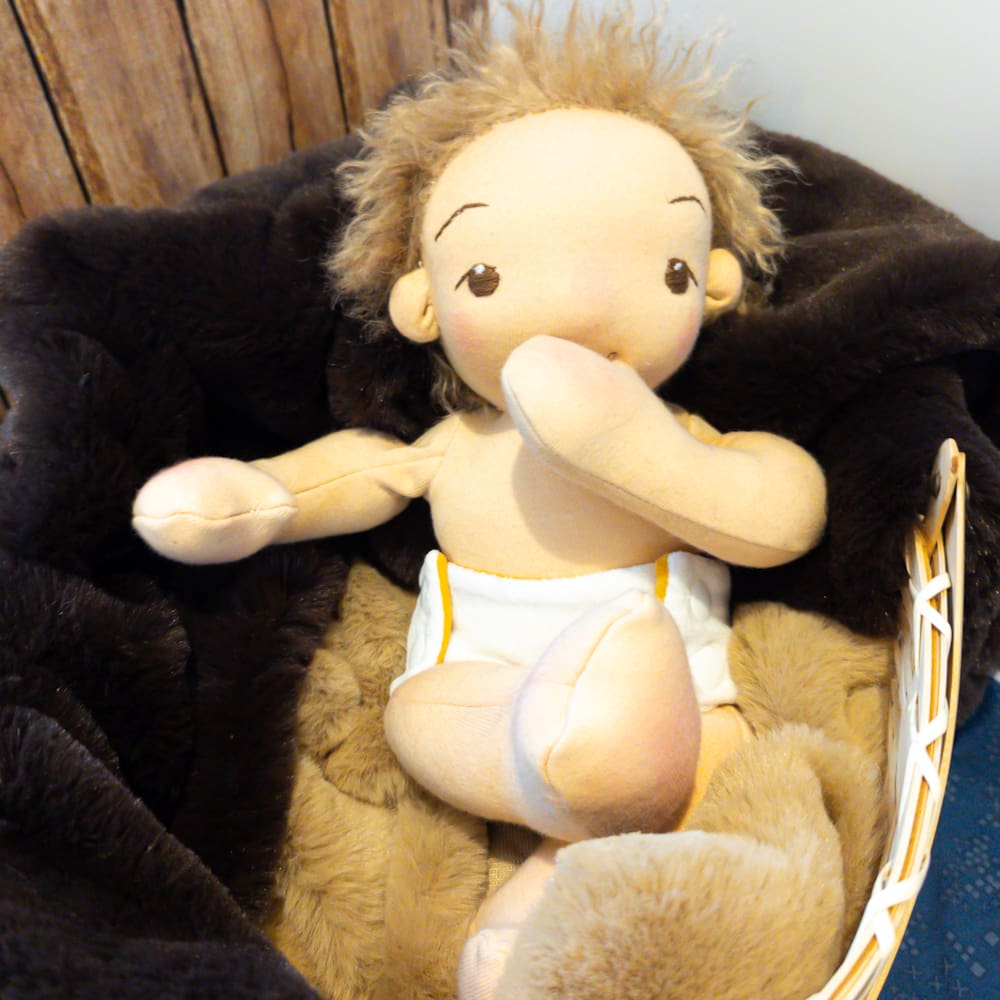 handmade fabric baby doll with sandy brown hair and brown eyes lying in a basket dressed in a white nappy with his arm raised to his mouth
