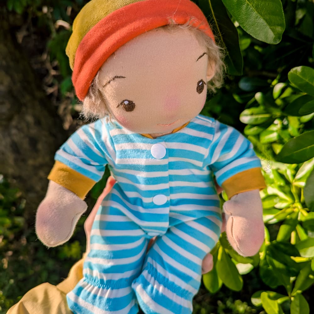 handmade fabric baby doll with light brown hair and brown eyes wearing a hat and a a striped onesie sitting on a hand with a garden backdrop