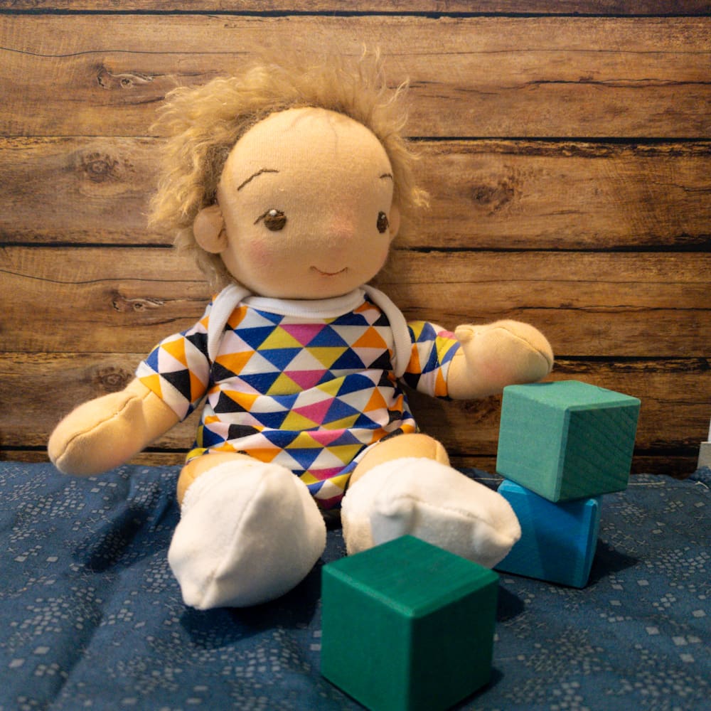 handmade fabric baby doll with light brown hair pale skin and brown eyes sitting playing with bricks in front of a plank wood background