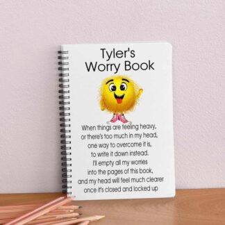 A spiral-bound A5 notebook on a wooden desk. The cover features a fuzzy yellow monster in red sneakers and a poem about writing down worries to clear the mind. The title reads "Tyler's Worry Book" at the top.
