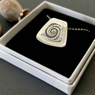 A square silver pendant with a fossil embossed on the front resting on a small white gift box.