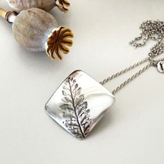 A fine silver clay square pendant with a lean impression that has been patinerd in the centre. It has a sterling silver chain and is displayed on a white washed wood background.