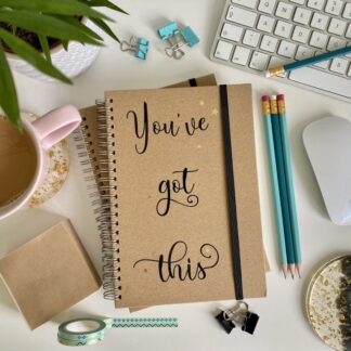 A desk with a cup of tea, pencils, keyboard & mouse and other stationery items. In the centre of the desk is an A5 notebook. The notebook has the text 'You've Got This' in black script style font.