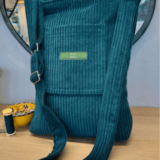 The photograph shows an aqua green corduroy handbag resting on a table with a mirror behind it. The handbag has a long strap attached with a silver adjustable slider on it. To the side of the handbag is a small pot of needle pins and a reel of cotton in the same aqua green colour as the handbag