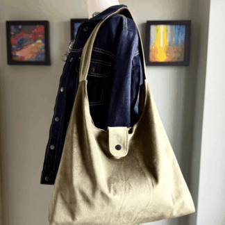 The photograph shows an olive green velvet bag resting on the shoulder of a dressed mannequin. The mannequin is wearing a navy blue denim jacket. In the background are 3 colourful small abstract prints