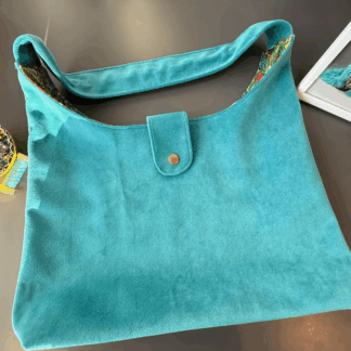 The photograph shows a turquoise velvet hobo bag rested against a grey desk. To the left of the bag is a yellow pot of needle pins and a reel of turquoise thread. To the right of the bag is a section of a photograph showing a turquoise feather long coat