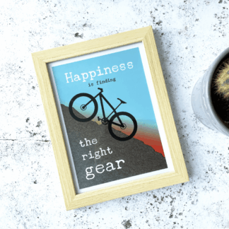 A photograph on an A5 motivational print framed and taken at an angle. The print has text that says: "Happiness is finding the right gear" as a mountain bike rides up a steep hill as the sun sets behind.