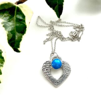 A fine silver heart pendant which has a textured pattern and a rich blue cabochon and a sterling silver chain. It is displayed on a white back ground with green ivy leaves in the left top corner.
