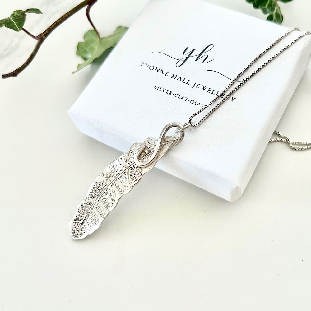 A fine silver pendant in the shape of a feather. Displayed sat on a white gift box.