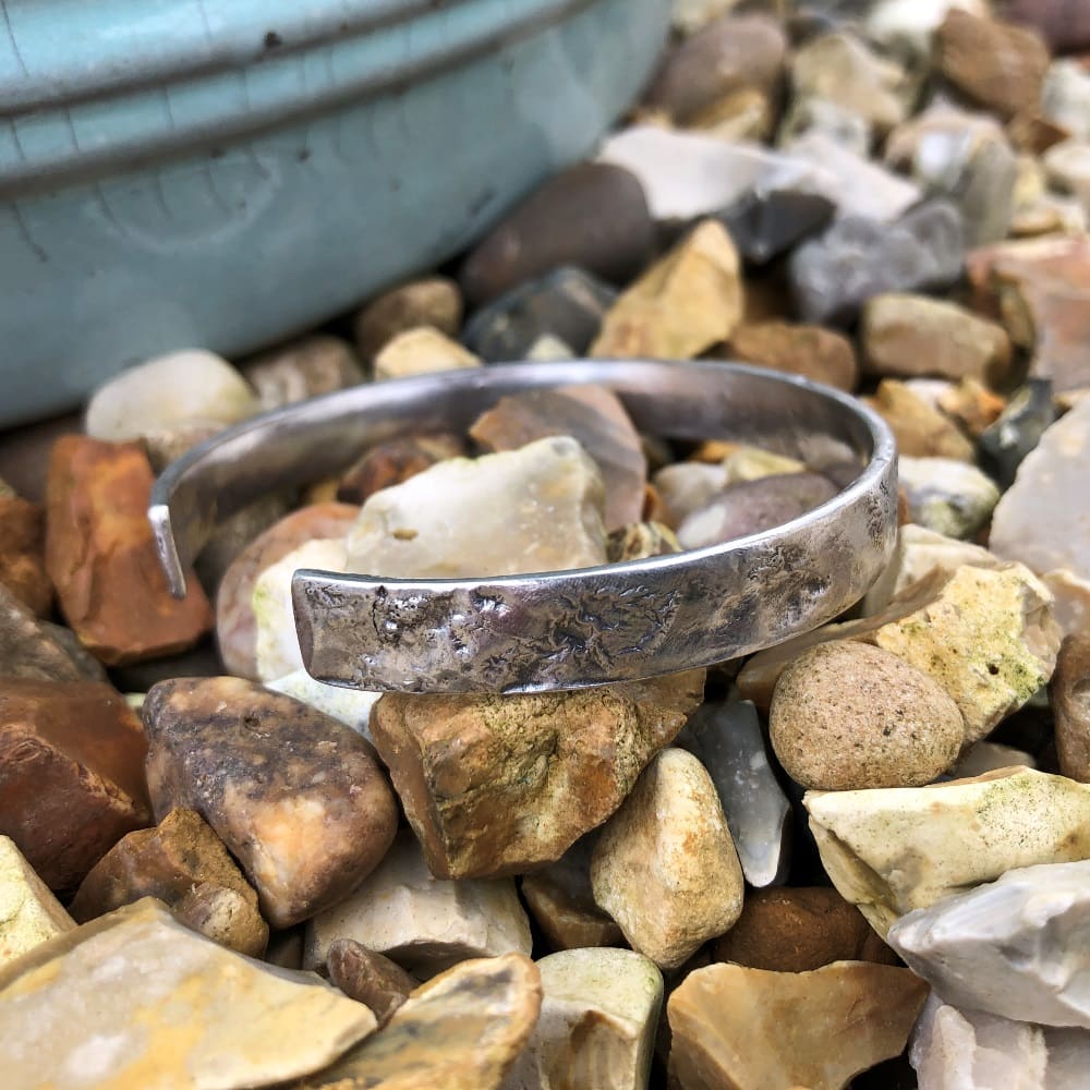 A sustainable textured silver cuff bangle lying on pebbles, highlighting its coastal inspiration.
