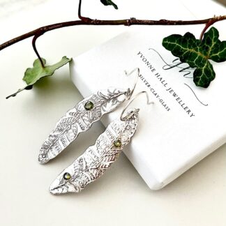 A pair of fine silver drop earrings in the shape of feather with small 5mm. peridot cubic zirconia stones. They are displayed leasing against a white presentation box with ivy leaves to decorate.