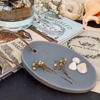 Coast grey soy wax melt and wardrobe freshener with small white shells and dried white flower embeds sits on a book next to the Coast botanical candle surrounded by shells.