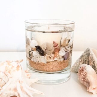 Coast botanical candle with natural coloured sand and shells rests on a surface between two large seashells