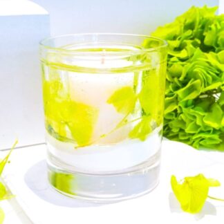 Fresh and vibrant green grass and hydrangea scented gel and soy wax botanical candle decorated with vibrant green real hydrangea petals is placed on a pure white surface next to a bright green hydrangea flower.