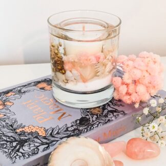 Pink champagne soy and gel wax candle decorated with pastel pink and white flowers sits on top of a grey book next to sea shells, pink dried flowers and pieces of rose quartz.