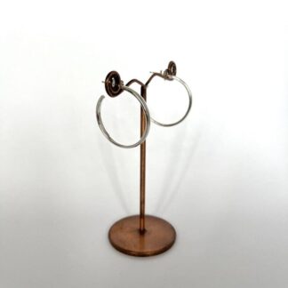 A pair of handmade sterling silver hoop earrings on a display stand. They are a medium size and they are on a white background. They have a post and scroll fitting for the ear lobe