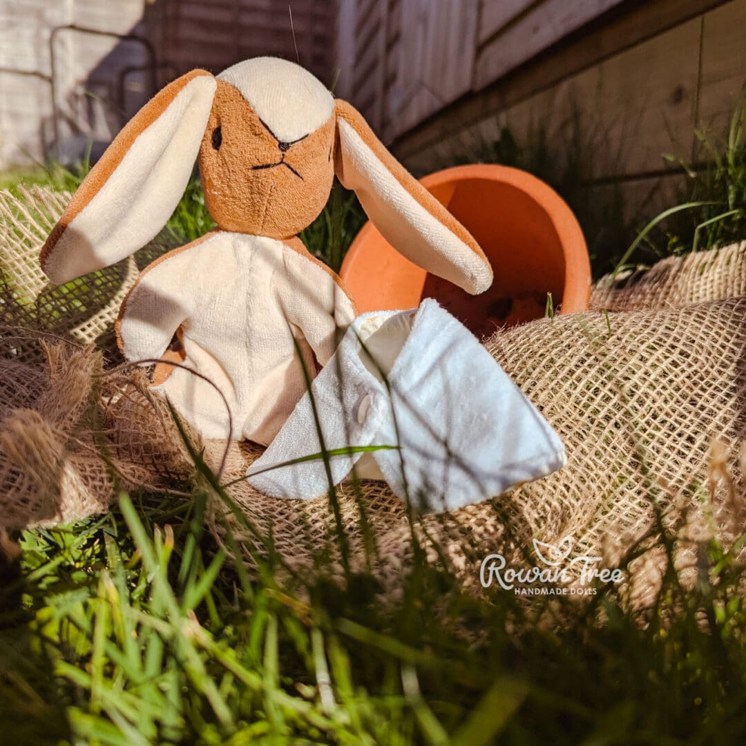 handmade velveteen bunny in caramel and cream colours with light blue cloak next to her sat in front of plant pot