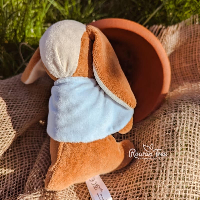 Caramel velveteen bunny from the rear looking into a plant pot wearing a light blue cape