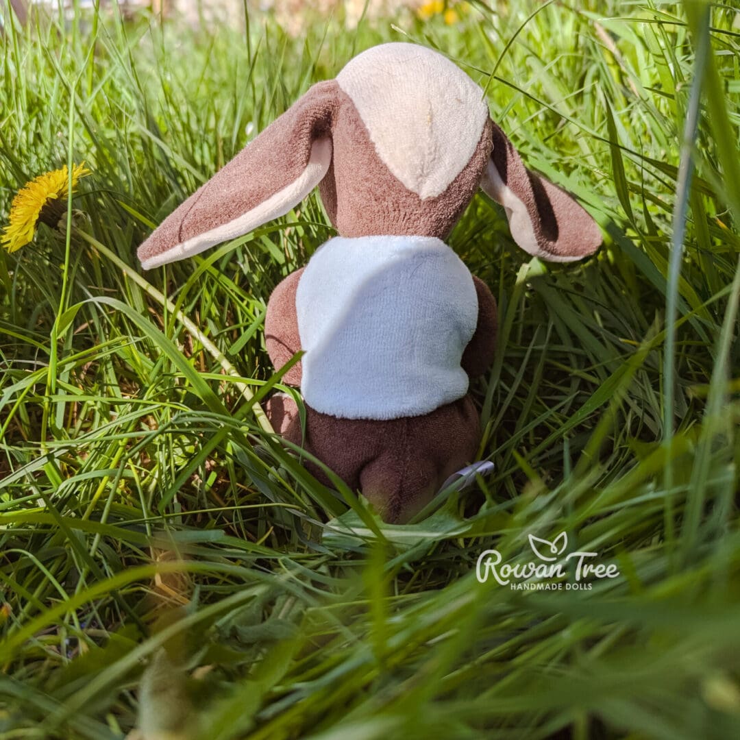 Handmade chocolate and cream furred velveteen bunny seen from rear wearing light blue velveteen double breasted waistcoat sat on hessian on grass