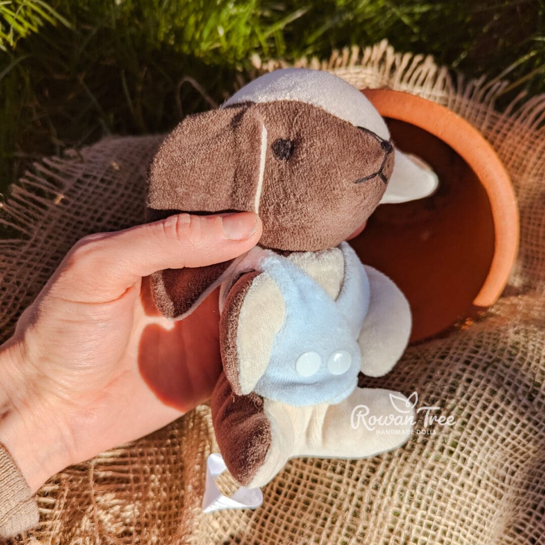 Handmade chocolate and cream furred velveteen bunny wearing light blue velveteen double breasted waistcoat held in maker's hand in garden