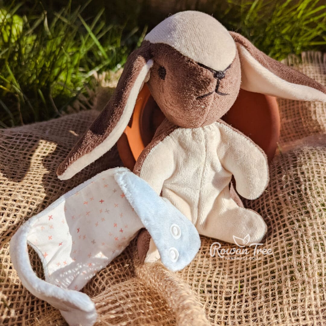 Handmade chocolate and cream furred velveteen bunny next to his light blue velveteen double breasted waistcoat sat on hessian on grass