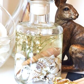 Coast luxury botanical diffuser. Clear fresh scented fragrance oil in a 200ml apothecary bottle with shells and small white dried flowers suspended in the liquid. The reed diffuser is placed on a white surface with a large white flower and a bronze hare ornament.