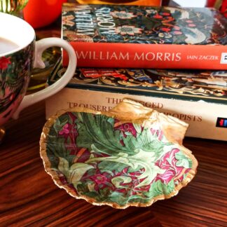 Handcrafted gilded, decoupaged scallop shell trinket dish, decorated with the William Morris Granville design in green and dark red. The shell dish is placed on a coffee table next to a china cup and two books.