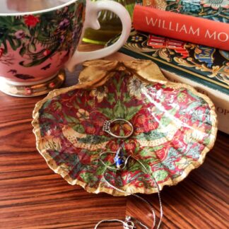 Large gilded scallop shell trinket dish decorated with The strawberry Thief design on a deep red background. The dish is on a coffee table next to books and a china cup.