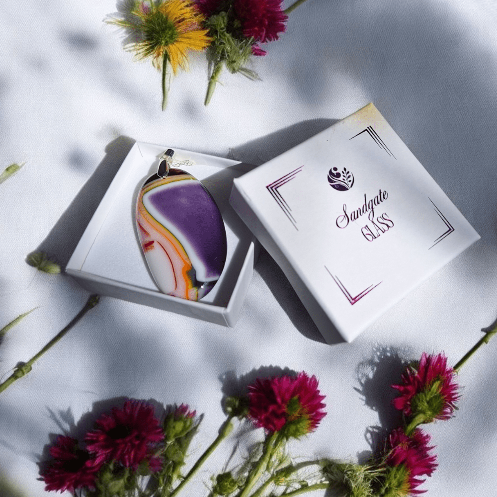 a handmade fused glass pendant in purple, white, and amber with a silver-plated bail and chain, photographed beside wildflowers in natural light.