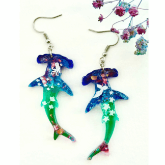 A pair of resin hammerhead shark earrings in blue and green gradient, filled with glitter and miniature ocean creatures like turtles, whales, and octopuses, displayed with pink glitter shimmer in the tail.
