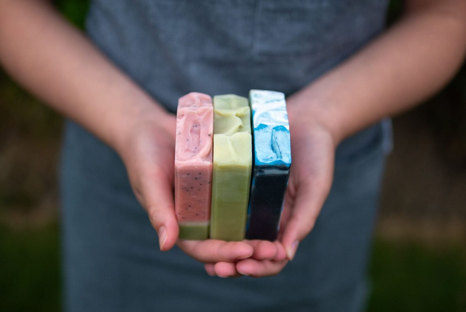 Person holding four handmade soap bars in natural colours — pink, green, cream, and blue — outdoors.