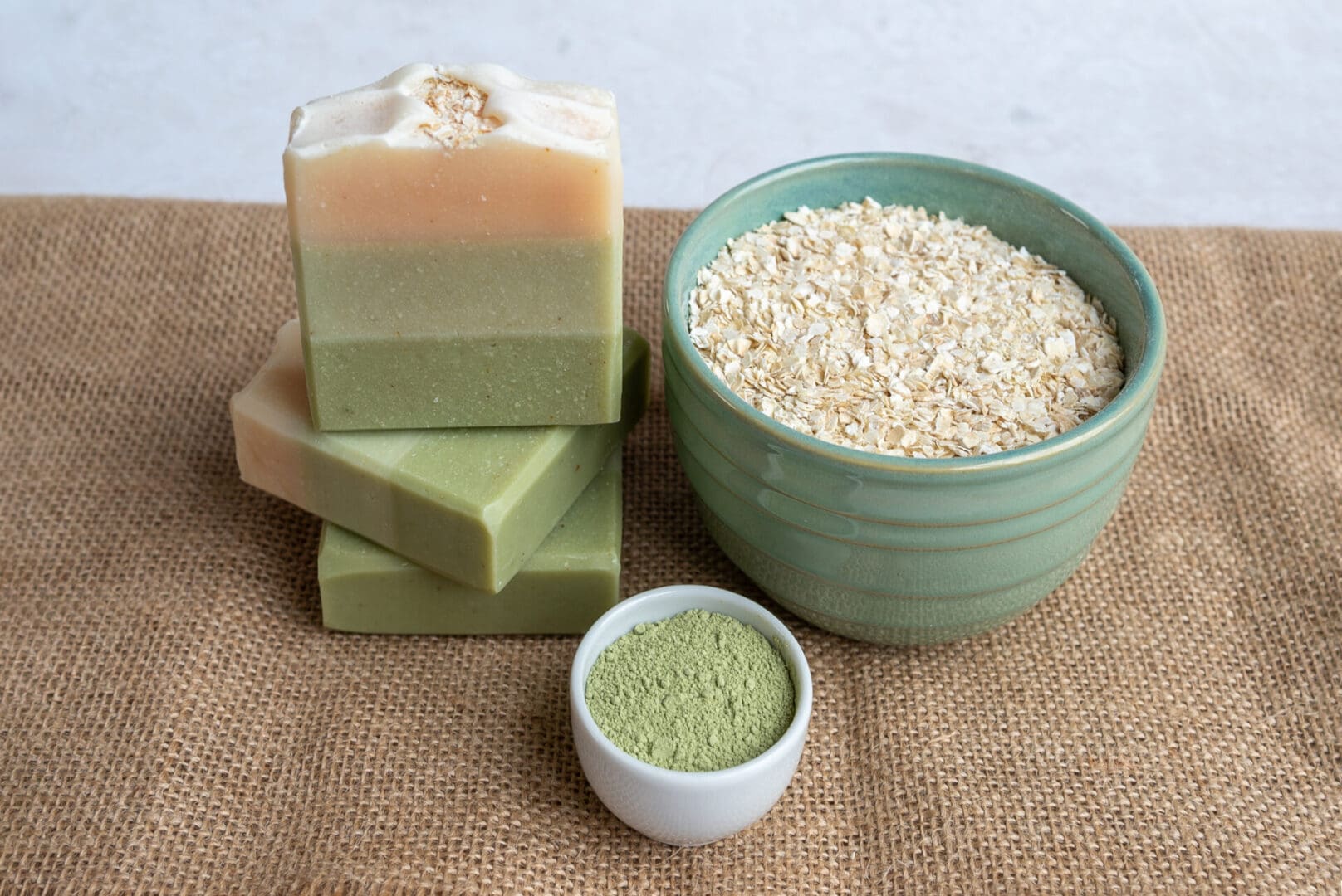 Green layered soap bars sitting on a brown cloth with a bowl of oats and a bowl of green clay