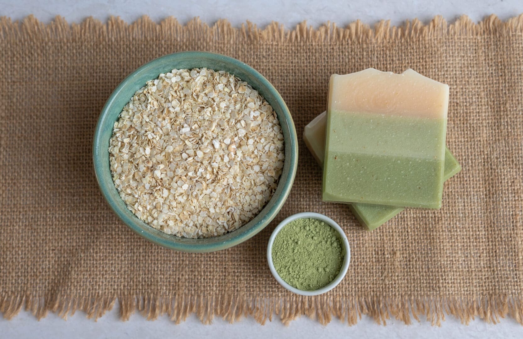 Green layered soap bars sitting on a brown cloth with a bowl of oats and a bowl of green clay