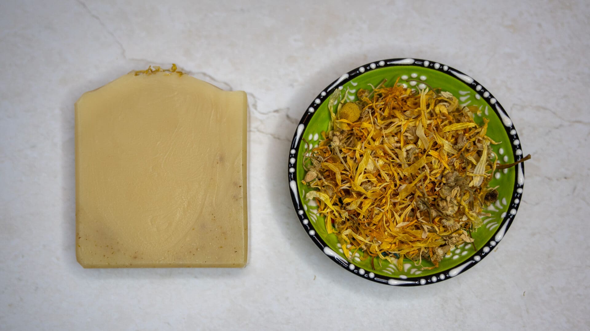 Yellow, natural soap bar next to a bowl of yellow calendula and chamomile flower petals