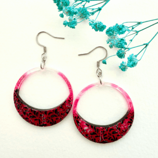 The image shows a round hoop earring on an earring hook. The earring features an enlarged lower section with a pink lace/leaf pattern with a black marble like background with white veins. Pink spreads up the sides of the hoop.