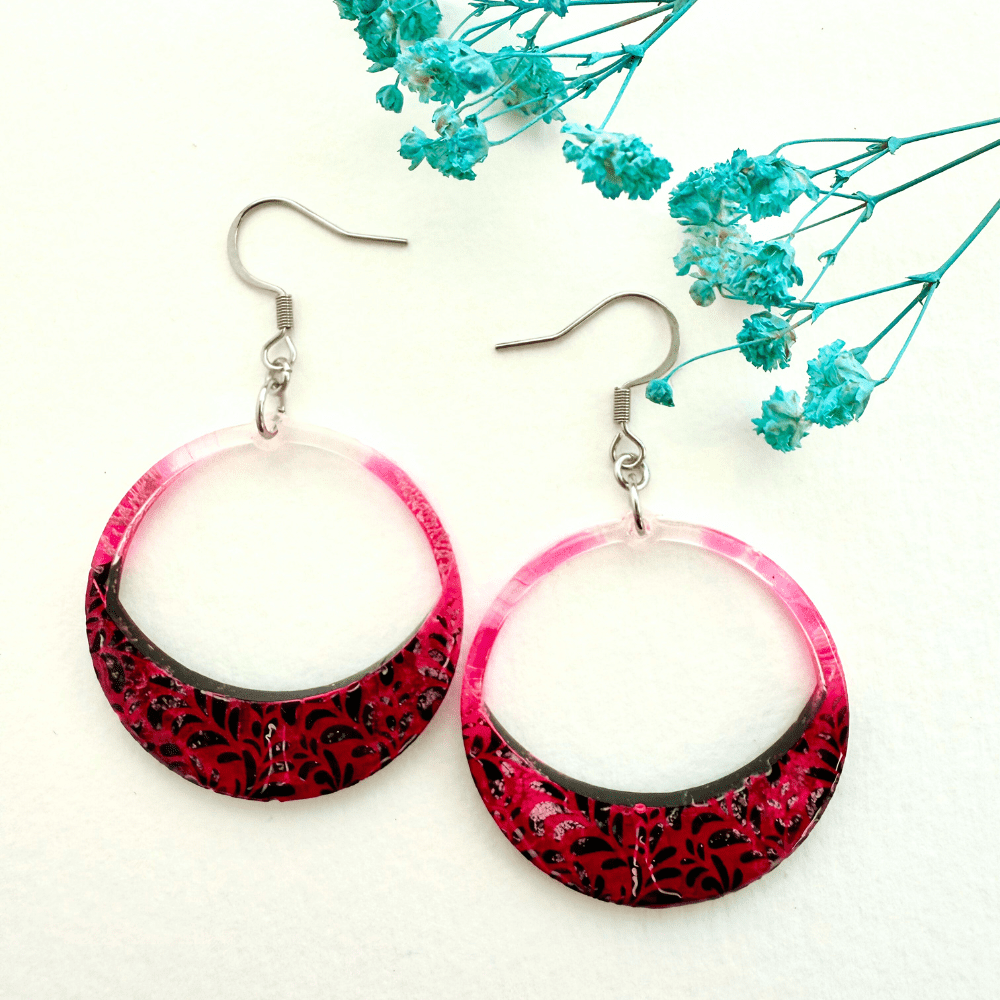 The image shows a round hoop earring on an earring hook. The earring features an enlarged lower section with a pink lace/leaf pattern with a black marble like background with white veins. Pink spreads up the sides of the hoop.